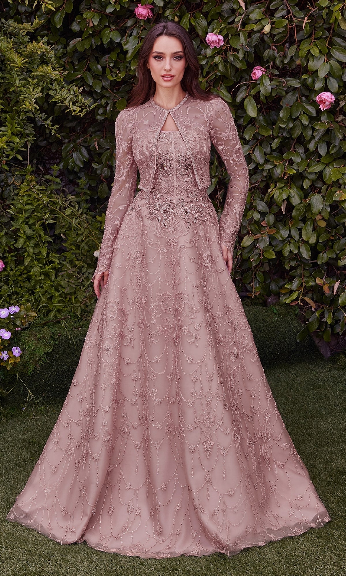 Woman wearing the Andrea & Leo MA104 Long Sleeve Long Formal Dress, featuring blush pink embroidered lace and beaded details, standing outdoors among leafy greenery and pink flowers.